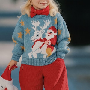 May include: A child in a light blue Christmas jumper featuring Santa Claus and a reindeer, paired with red corduroy trousers and a red beret. The jumper has yellow star details. The child holds a white stuffed toy with a red hat.