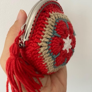 Pode incluir: Uma carteira de moedas de croch&ecirc; vermelha feita &agrave; m&atilde;o com um fecho prateado e uma borla vermelha a condizer. A carteira apresenta um design floral em bege, verde-azulado e branco. A carteira &eacute; segurada em uma m&atilde;o.