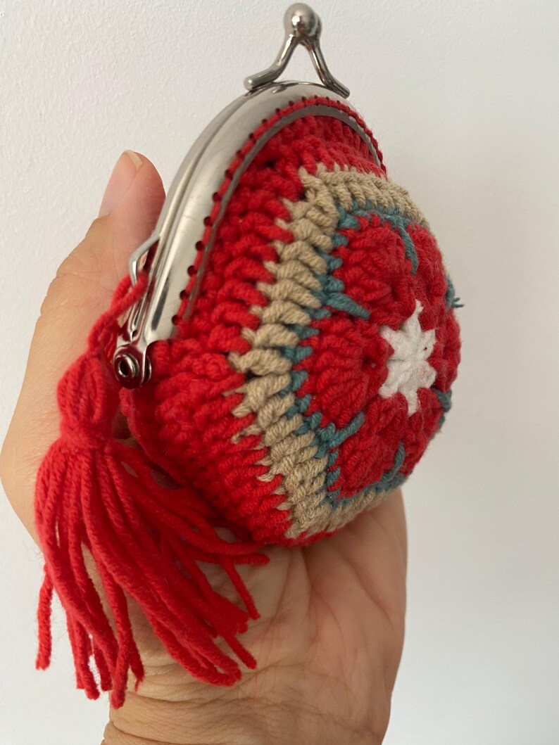 Pode incluir: Uma carteira de moedas de croch&ecirc; vermelha feita &agrave; m&atilde;o com um fecho prateado e uma borla vermelha a condizer. A carteira apresenta um design floral em bege, verde-azulado e branco. A carteira &eacute; segurada em uma m&atilde;o.