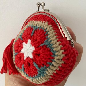 Pode incluir: Uma carteira de moedas de croch&ecirc; feita &agrave; m&atilde;o em vermelho, com um design floral em branco, bege e azul-petr&oacute;leo. A carteira tem um fecho prateado e uma borla vermelha. A carteira &eacute; segurada em uma m&atilde;o.