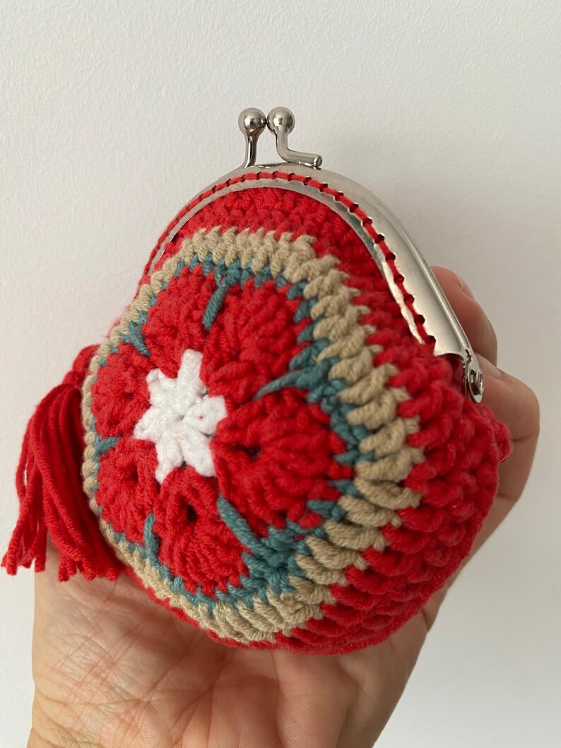 Pode incluir: Uma carteira de moedas de croch&ecirc; feita &agrave; m&atilde;o em vermelho, com um design floral em branco, bege e azul-petr&oacute;leo. A carteira tem um fecho prateado e uma borla vermelha. A carteira &eacute; segurada em uma m&atilde;o.
