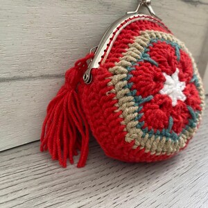 Pode incluir: Uma carteira redonda de croch&ecirc; vermelha feita &agrave; m&atilde;o, com fecho prateado e borla a condizer. O design circular apresenta detalhes em bege, turquesa e branco. O trabalho de croch&ecirc; &eacute; detalhado e &eacute; um acess&oacute;rio &uacute;nico.