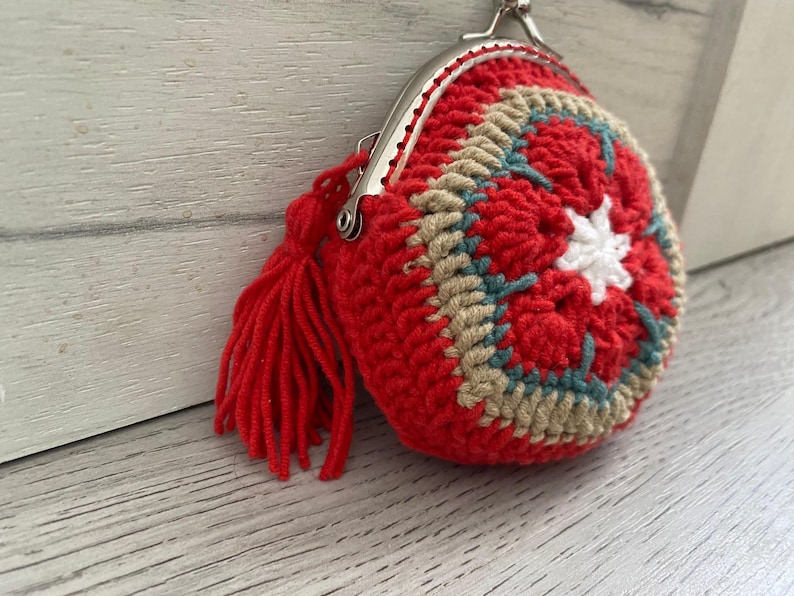 Pode incluir: Uma carteira redonda de croch&ecirc; vermelha feita &agrave; m&atilde;o, com fecho prateado e borla a condizer. O design circular apresenta detalhes em bege, turquesa e branco. O trabalho de croch&ecirc; &eacute; detalhado e &eacute; um acess&oacute;rio &uacute;nico.