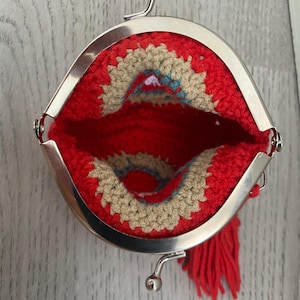 Pode incluir: Uma bolsa de moedas vermelha de croch&ecirc; com fecho prateado. O interior apresenta um design bege e turquesa. Uma borla vermelha pende da lateral. A bolsa est&aacute; aberta, mostrando os detalhes internos.