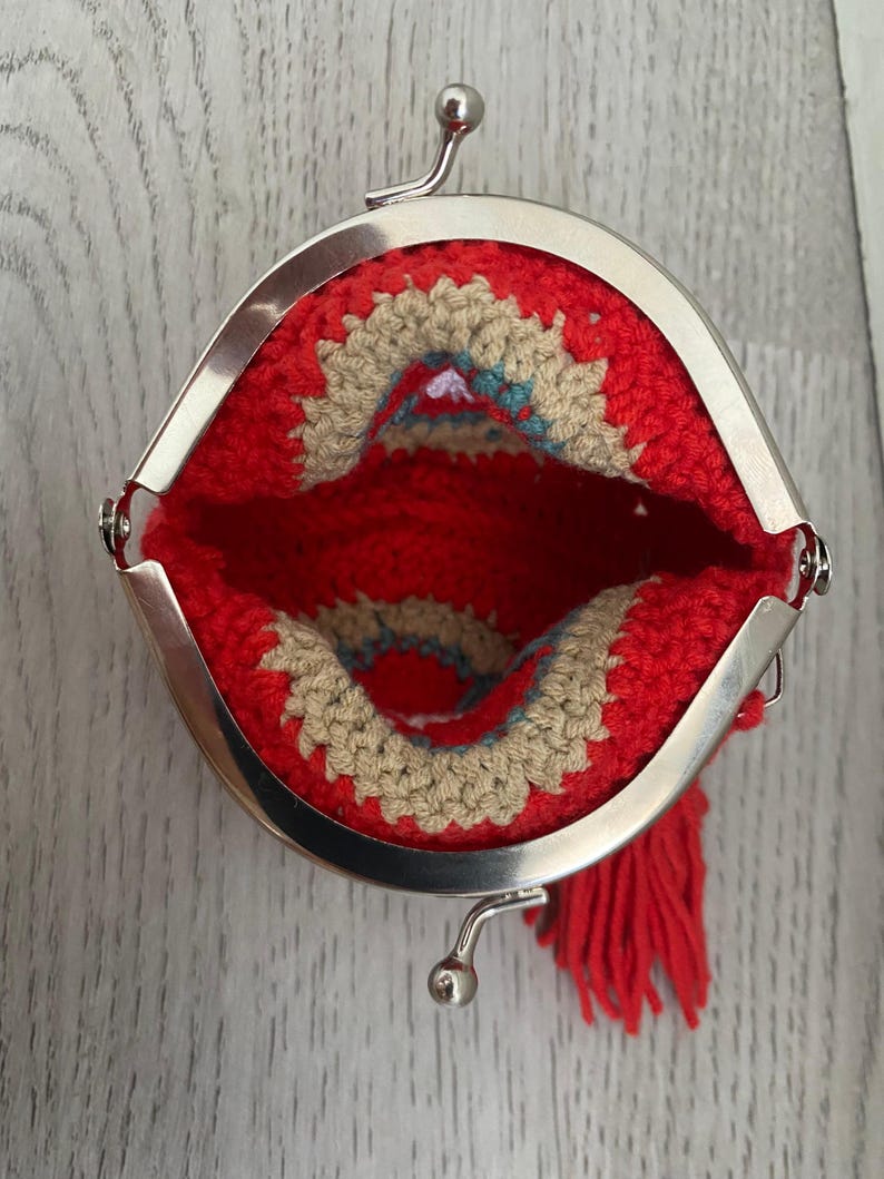 Pode incluir: Uma bolsa de moedas vermelha de croch&ecirc; com fecho prateado. O interior apresenta um design bege e turquesa. Uma borla vermelha pende da lateral. A bolsa est&aacute; aberta, mostrando os detalhes internos.