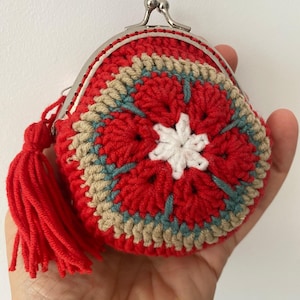 Pode incluir: Uma carteira de moedas redonda de croch&ecirc; vermelha com um padr&atilde;o floral em branco, bege e azul. Possui um fecho prateado e uma borla vermelha. A carteira &eacute; feita &agrave; m&atilde;o. A carteira &eacute; segurada em uma m&atilde;o.