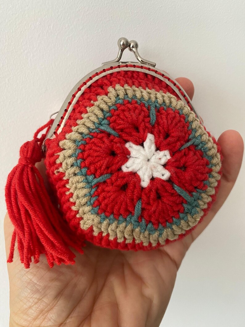 Pode incluir: Uma carteira de moedas redonda de croch&ecirc; vermelha com um padr&atilde;o floral em branco, bege e azul. Possui um fecho prateado e uma borla vermelha. A carteira &eacute; feita &agrave; m&atilde;o. A carteira &eacute; segurada em uma m&atilde;o.