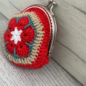 Pode incluir: Uma carteira de moedas de croch&ecirc; feita &agrave; m&atilde;o em vermelho, bege e azul-petr&oacute;leo. A carteira apresenta um design de estrela na frente e um fecho de metal prateado. O trabalho de croch&ecirc; &eacute; detalhado e a carteira &eacute; pequena e redonda.