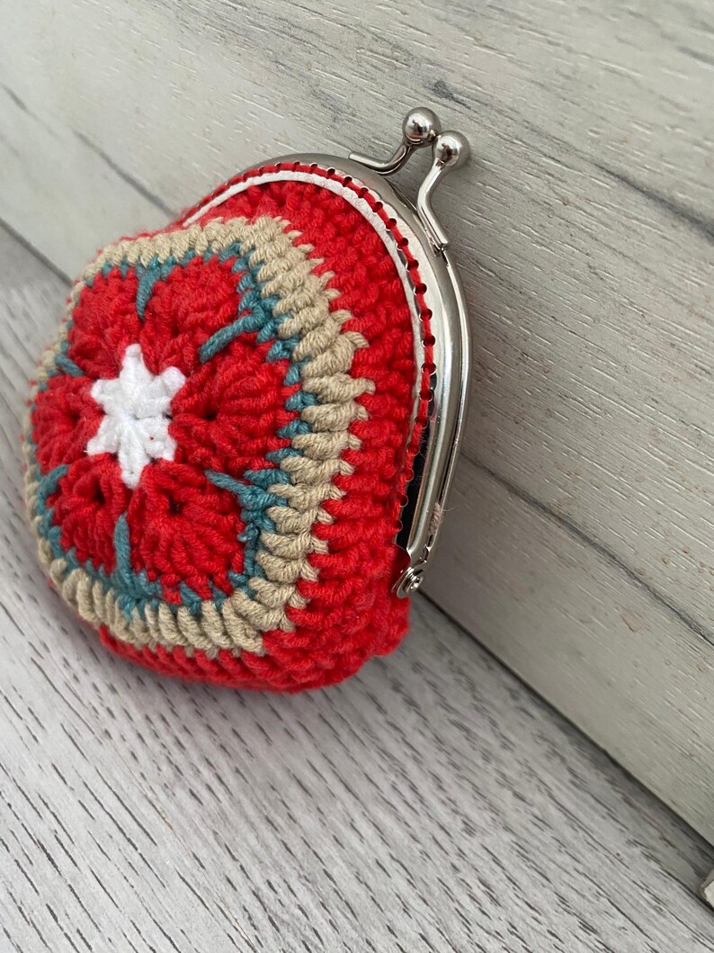 Pode incluir: Uma carteira de moedas de croch&ecirc; feita &agrave; m&atilde;o em vermelho, bege e azul-petr&oacute;leo. A carteira apresenta um design de estrela na frente e um fecho de metal prateado. O trabalho de croch&ecirc; &eacute; detalhado e a carteira &eacute; pequena e redonda.