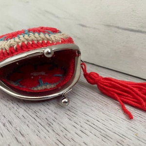 Pode incluir: Uma carteira de moedas vermelha de croch&ecirc; com fecho prateado e borla vermelha a condizer. A carteira tem um design circular com detalhes em vermelho, bege e azul. O interior &eacute; vermelho com um design azul e bege.