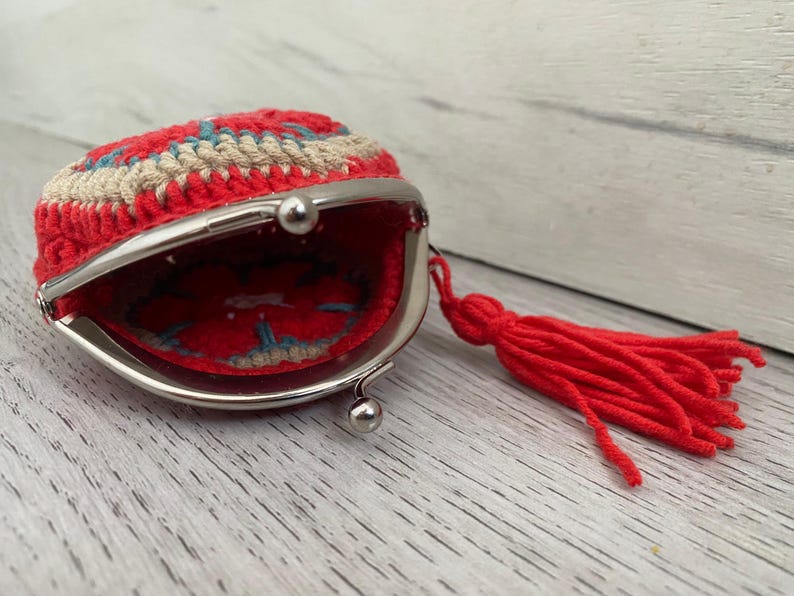 Pode incluir: Uma carteira de moedas vermelha de croch&ecirc; com fecho prateado e borla vermelha a condizer. A carteira tem um design circular com detalhes em vermelho, bege e azul. O interior &eacute; vermelho com um design azul e bege.