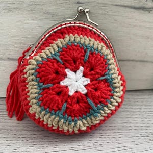 Pode incluir: Uma carteira de moedas de croch&ecirc; feita &agrave; m&atilde;o em vermelho, bege, azul-petr&oacute;leo e branco. A carteira redonda apresenta um design floral e um fecho de metal. Uma borla vermelha pende de um lado. A carteira est&aacute; em uma superf&iacute;cie de madeira clara.