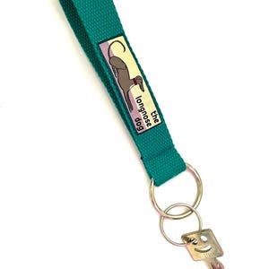 May include: Teal key wrist strap with a metal key ring and a key. The strap has the text "the longnose dog" and a graphic of a greyhound dog. The key has a smiley face.