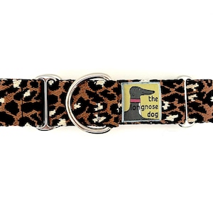 May include: A dog collar with a brown and black leopard print pattern. The collar has a silver metal D-ring and hardware. A small tag features a greyhound illustration and the text "the longnose dog".