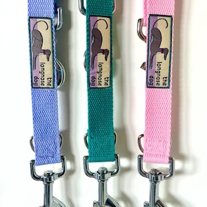 May include: Three dog leashes in light purple, teal, and light pink. Each leash features a woven fabric strap, a silver metal swivel snap hook, and a rectangular fabric label with an illustration of a dog and the text the longnose dog.