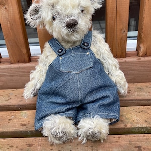 May include: A handmade teddy bear with cream-colored fur and dark accents on the ears and nose. The bear is wearing blue denim overalls with metal button closures. The bear is sitting on a wooden bench.