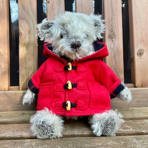 Puede incluir: Un oso de peluche hecho a mano con pelaje gris claro, que lleva un abrigo tipo duffel rojo brillante con cierres de palanca negros y un forro a cuadros. El oso está sentado en un banco de madera, mostrando su detallada artesanía.