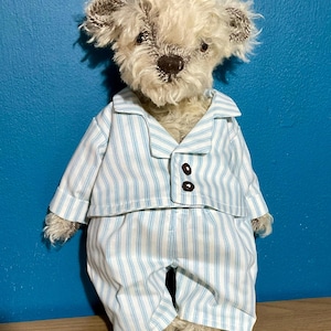 May include: A teddy bear wearing blue and white striped pyjamas. The bear has a light brown nose and ears, and the pyjamas have two brown buttons. The background is a teal colour.