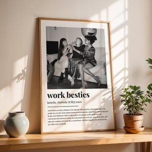 May include: Black and white framed print featuring three people celebrating. The print includes the text "work besties" and a definition. The frame is a light brown color, and the print is displayed on a wooden surface.
