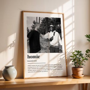 May include: A framed black and white photograph featuring two people, with the word "homie" and its definition below. The photograph is in a wooden frame and is displayed on a wooden surface.