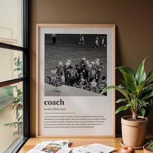 May include: A framed black and white photograph of a football team huddle. The word "coach" is printed in a bold font at the bottom, along with a definition. The frame is light-colored, and the image is displayed on a wooden surface.