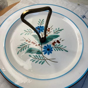 May include: White ceramic serving plate with a blue rim and a metal handle. The plate features a floral design with blue flowers, green leaves, and brown berries. The handle is a dark, aged metal.