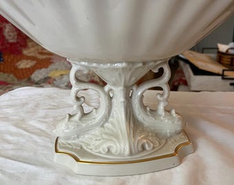 May include: A white porcelain bowl with a scalloped edge and gold trim sits on a decorative stand with a gold trim. The stand features a floral design and two dolphin figures.