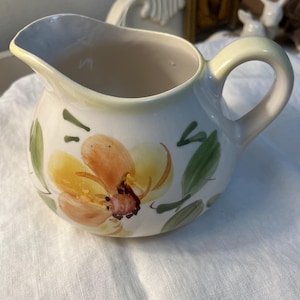 May include: Cream-colored ceramic creamer with a yellow and orange floral design and green leaves. The creamer has a curved handle and a spout for easy pouring. The floral design is hand-painted, adding a unique touch to the piece.