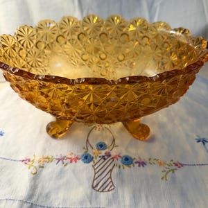 May include: Amber-colored, footed glass bowl with a scalloped rim. The bowl features a geometric pattern and rests on three small feet. The bowl is likely vintage and suitable for serving fruit or displaying decorative items.