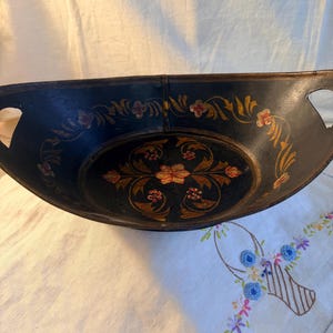 May include: A black oval bowl with integrated side handles, featuring hand-painted floral motifs in gold, red, and green. The interior base displays a symmetrical flower design, while the curved sides are decorated with trailing leaf patterns.