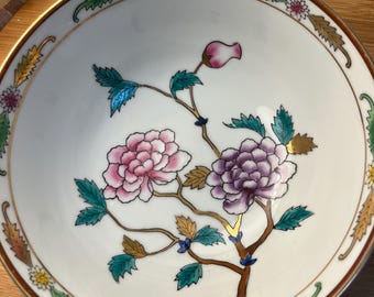 Puede incluir: Un cuenco de cerámica blanca con un diseño floral. El cuenco presenta flores rosas y moradas con hojas verdes y detalles dorados. El borde tiene un borde marrón con un patrón decorativo. El cuenco está sobre una superficie de madera.