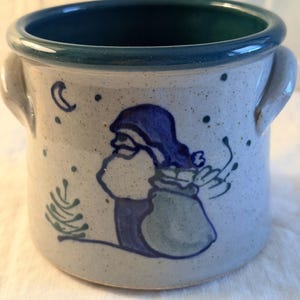 May include: A ceramic pot with a blue rim and handles. The pot is decorated with a blue Santa Claus design carrying a sack, a crescent moon, a Christmas tree, and dots. The pot has a speckled, off-white background.