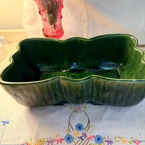 May include: A dark green, leaf-shaped ceramic dish with a glossy finish. The dish is set on a white embroidered tablecloth with a floral design. A pink glass vase with flowers is in the background.