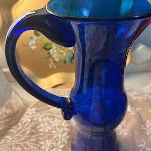May include: A cobalt blue glass pitcher with a large handle and a flared rim. The pitcher has a bulbous base and a smooth, glossy finish. The background includes a gold-colored tray.