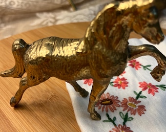 May include: A gold-colored metal horse figurine in a trotting pose. The horse is on a wooden surface, with a white cloth featuring embroidered red and pink flowers in the background. The figurine is approximately 5 inches long.