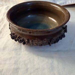 May include: A handmade ceramic bowl with a dark brown exterior and a blue-green interior. The rim has a textured, rough design. This unique bowl is suitable for serving food or as a decorative piece.