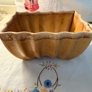 May include: A tan ceramic planter with a scalloped rim. The rectangular planter has a ribbed design and a light brown speckled rim. A pink glass vase is visible in the background.