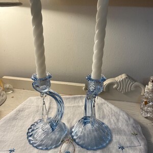 May include: Two blue glass candle holders with white spiral candles. The candle holders have a curved design with a crystal pendant. The base of the candle holders has a shell-like design. The candles are white.