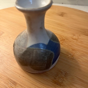 May include: A small, handcrafted ceramic vase with a rounded body and a flared neck. The vase features a color block design with shades of blue, gray, and brown. The interior is white, and the vase sits on a wooden surface.