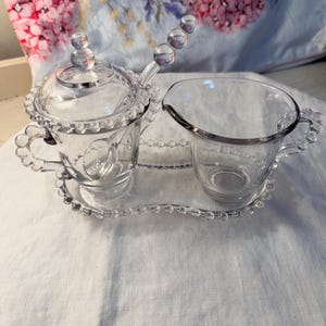 May include: A clear glass sugar and creamer set. The sugar bowl has a lid and a spoon with a decorative handle. The creamer has a spout. Both sit on a matching tray with a beaded edge. The set is designed for serving.
