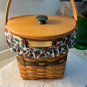 May include: A Longaberger Christmas Collection basket with a wooden lid and handle. The basket is woven with a light brown color and has a green trim. The lid is topped with a dark green knob and the basket is decorated with a fabric trim featuring red berries and green leaves.