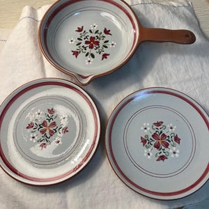 May include: A set of ceramic tableware featuring a floral design. The set includes a skillet with a handle and two plates. The plates and skillet have a light gray background with a red border and a floral pattern in the center.
