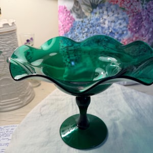 May include: A vibrant green glass pedestal bowl with a ruffled rim. The bowl sits on a short, dark green stem and a circular base. The glass has a glossy finish, reflecting light. The bowl is likely used for serving or display.