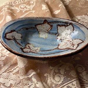 May include: Oval ceramic bowl with a blue interior and brown rim. The bowl features white leaf designs. The bowl is set on a tan lace tablecloth.