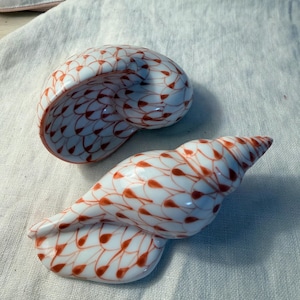 May include: Two white ceramic seashell figurines with a red teardrop pattern. One is a spiral shape, and the other is a conch shell shape. The seashells are on a white cloth surface.
