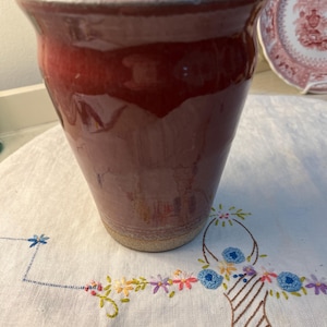 May include: A handcrafted, glazed ceramic vase in a deep red hue. The vase has a slightly flared rim and a smooth, glossy finish. The base is a lighter, unglazed color. The vase is sitting on a white embroidered cloth.