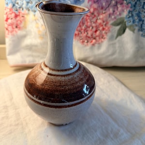 May include: A small, ceramic vase with a bulbous base and a flared top. The vase is primarily light gray with brown horizontal bands around the middle. The interior and rim are brown. The vase is sitting on a white cloth.