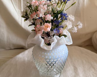May include: A clear glass vase with a hobnail texture and a ruffled white rim, filled with a bouquet of artificial flowers in shades of pink, blue, and white. The vase has a rounded body that tapers toward the base and a wide, wavy opening.
