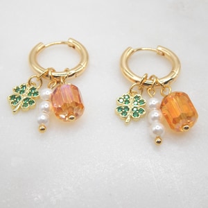 May include: Gold-coloured hoop earrings with hanging charms. Each earring has a green four-leaf clover, white pearls, and an orange crystal bead. The earrings are designed in a classic and elegant style.
