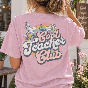 May include: A dusty pink short-sleeve t-shirt featuring a graphic on the back with the text "Cool Teacher Club" in large, bold, curved lettering. The design includes a white bird, colourful rainbow arcs, and white floral illustrations.